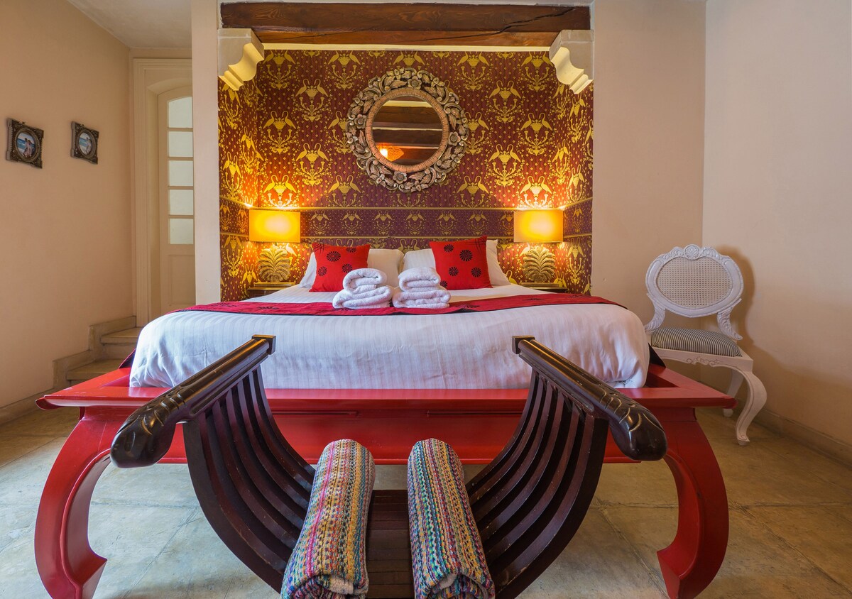 A spacious bedroom is characterized by a large bed adorned with white linens and red accent pillows. A round mirror is centered above the bed, reflecting warm lighting from bedside lamps. Colorful patterned decor and a decorative chair enhance the inviting environment.