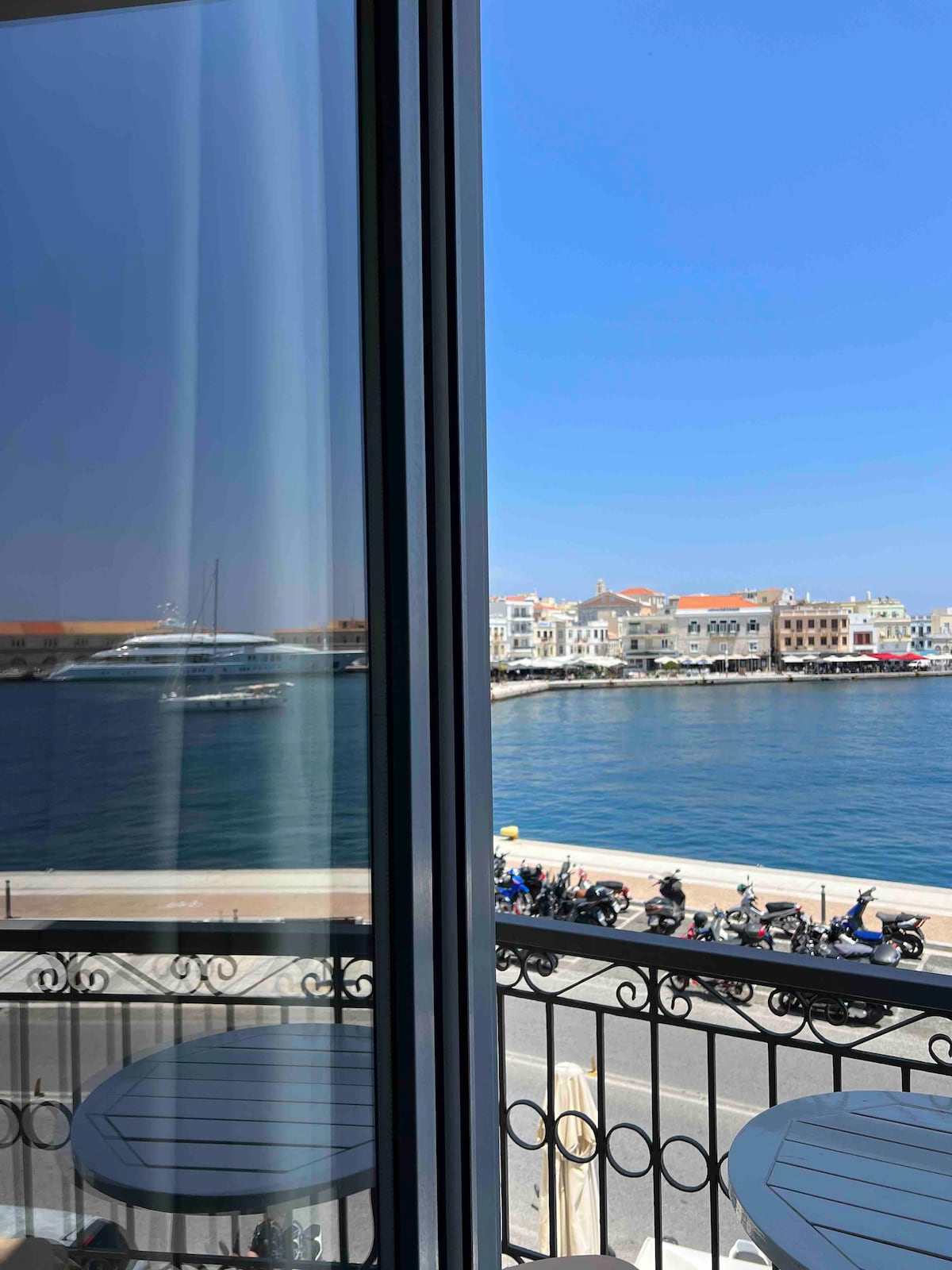 A view of the waterfront is framed by large glass doors, showing the harbor with boats docked and the city beyond. Scenic buildings line the shore, and parked scooters are visible along the promenade.