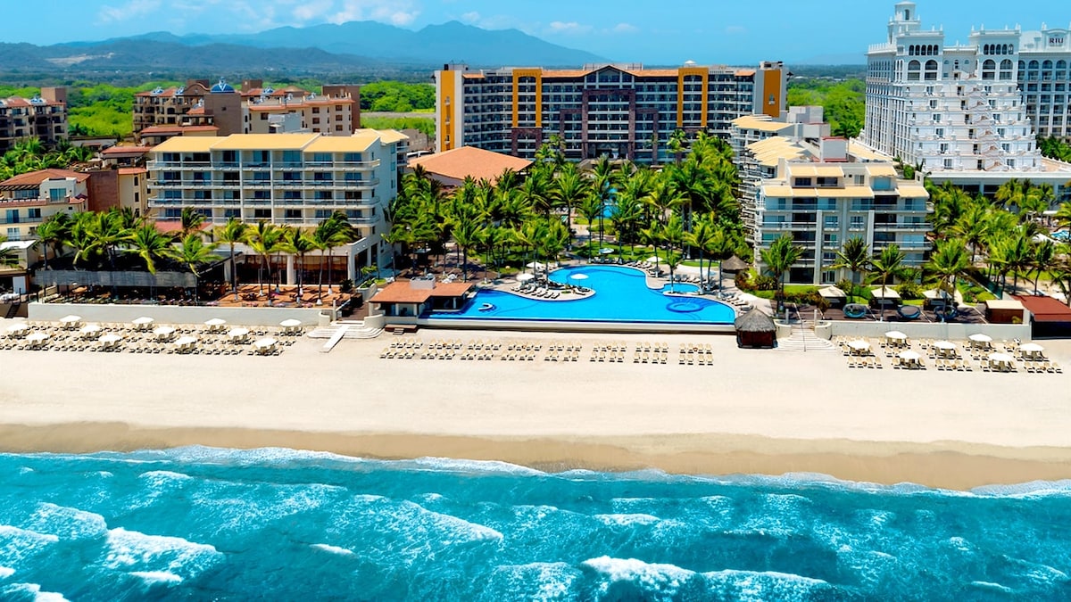 A beachfront resort is depicted, featuring a large swimming pool surrounded by palm trees and lounge chairs. The golden sand stretches along the shore, with waves gently lapping at the beach. In the background, several buildings provide a vibrant atmosphere against a clear blue sky.