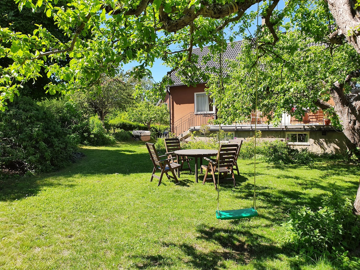 A spacious garden features a wooden swing hanging from a tree, accompanied by a table and several chairs. Lush green grass surrounds the seating area, and vibrant foliage adds to the natural setting. The house is visible in the background, complementing the peaceful atmosphere.
