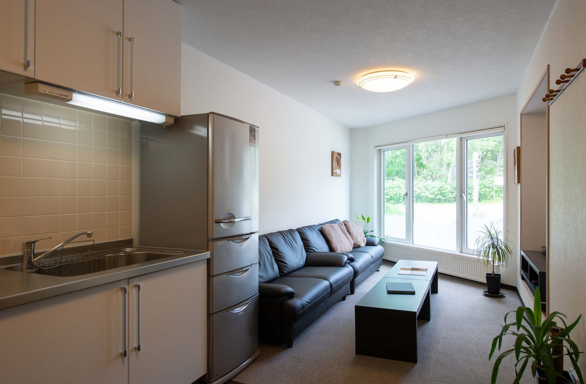 An open-plan living area is showcased, featuring a sleek kitchen with modern appliances. A comfortable black sofa lines one side, complemented by a coffee table. Large windows flood the space with natural light and offer views of greenery outside.
