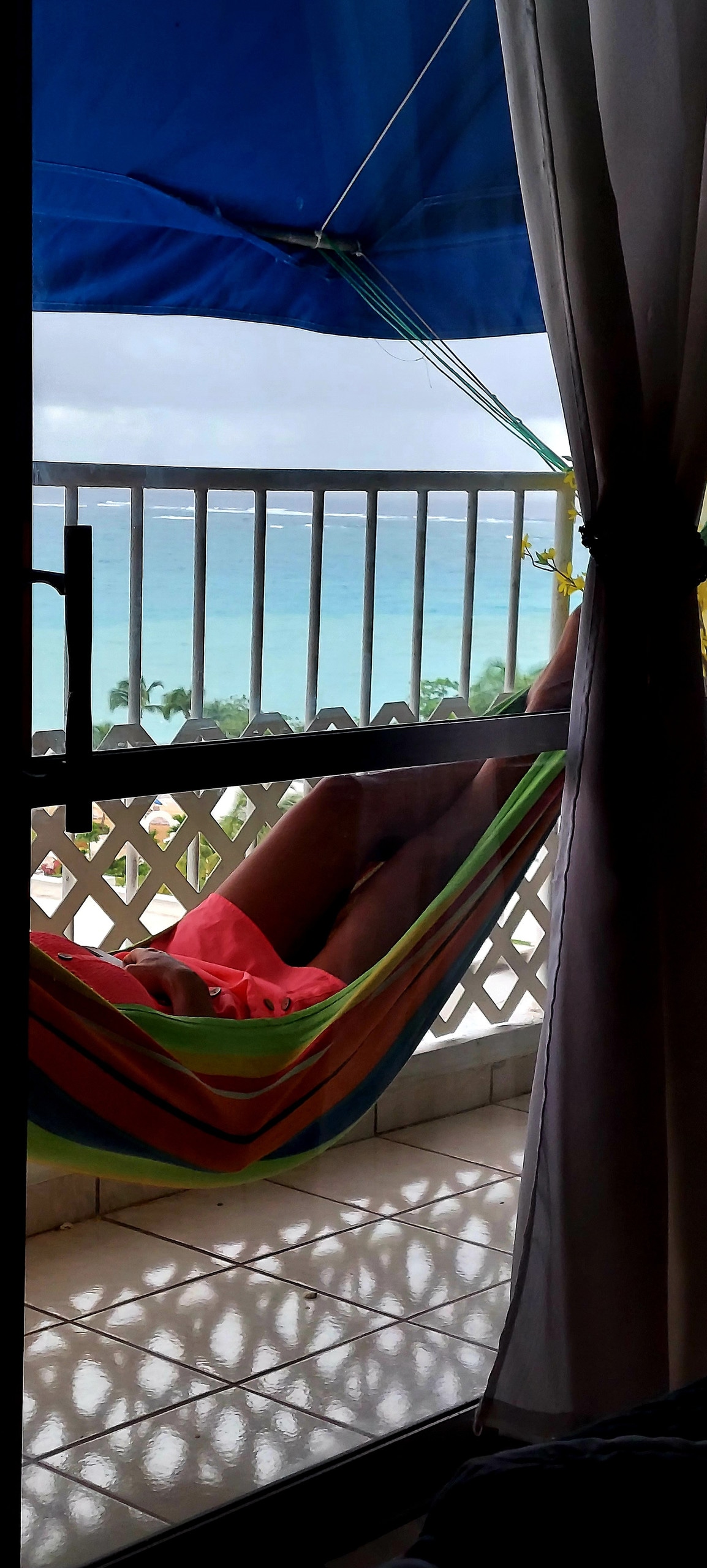 A colorful hammock is suspended on a balcony, with a view of the tranquil ocean in the background. Light blue water contrasts with the white waves, framed by a partially closed curtain. A pair of legs relax in the hammock, indicating a serene atmosphere.