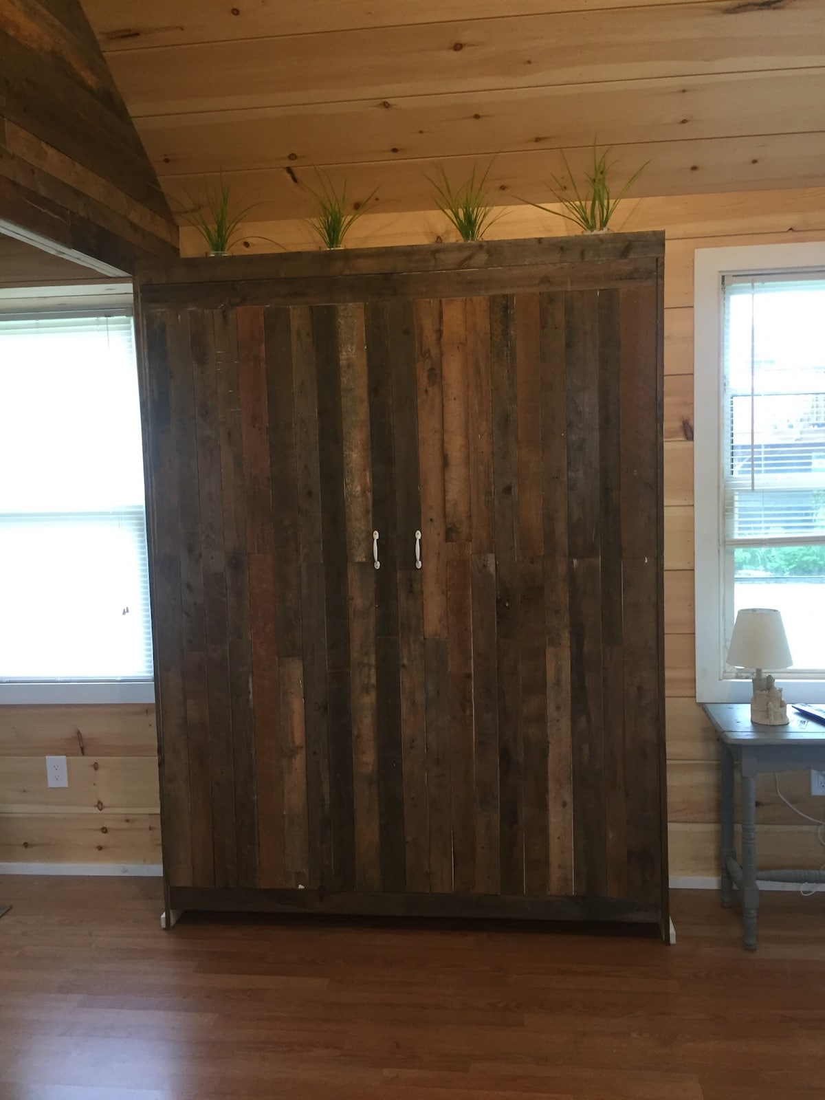 A rustic wooden wardrobe is featured, constructed from planks of varying shades of brown. It is positioned against a wall with ample natural light streaming in from two nearby windows. A small side table with a lamp is situated beside the wardrobe.