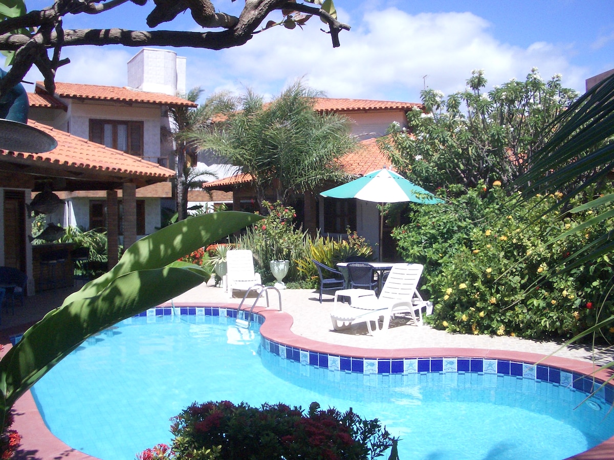 A serene swimming pool is surrounded by vibrant greenery and colorful flowers. Lounge chairs are positioned near the water, inviting relaxation. A shaded outdoor seating area appears to the left, adorned with plants and umbrellas, complementing the inviting ambiance of the villa.