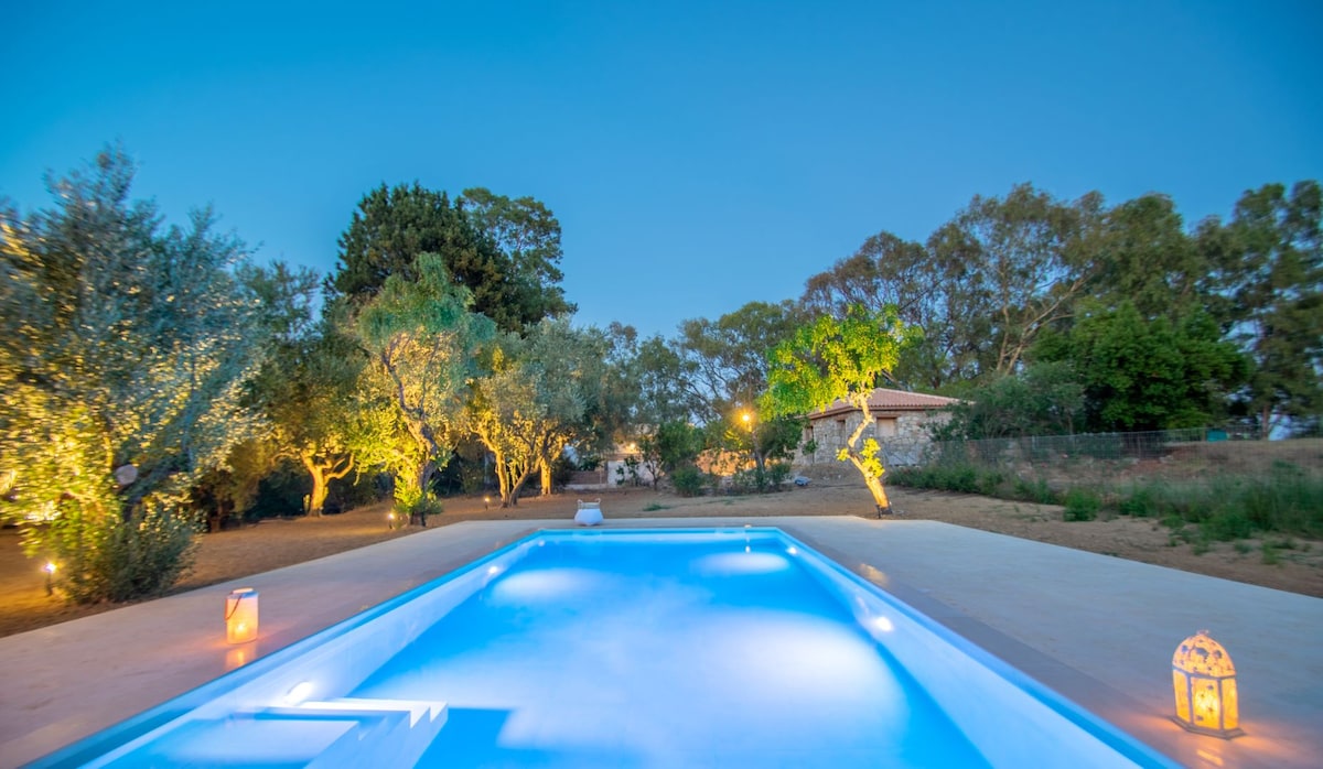 A private saltwater pool is illuminated with soft blue lighting, surrounded by olive trees and landscaped gardens. Lanterns cast a warm glow along the pool's edge, while the tranquil atmosphere is enhanced by the gentle lighting of nearby trees.