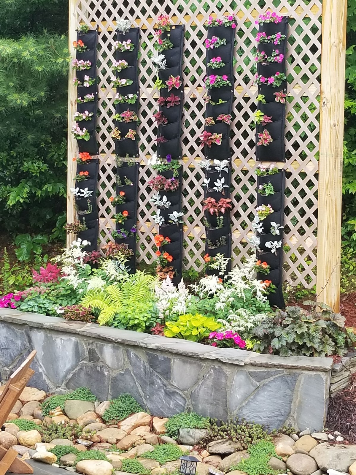 A vertical garden display is featured against a lattice backdrop, showcasing various colorful flowers and lush greenery. The arrangement includes multiple planters filled with vibrant blooms and a stone border surrounds a bed of low-growing plants, creating a vibrant natural setting.