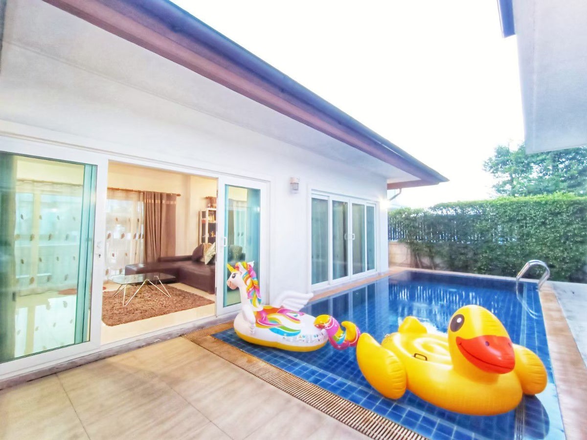 A private pool is featured, accompanied by colorful inflatable floats. Large glass doors allow natural light to enter the nearby living space, creating a seamless connection to the serene outdoor area. Lush greenery is visible beyond the pool, adding privacy to the setting.