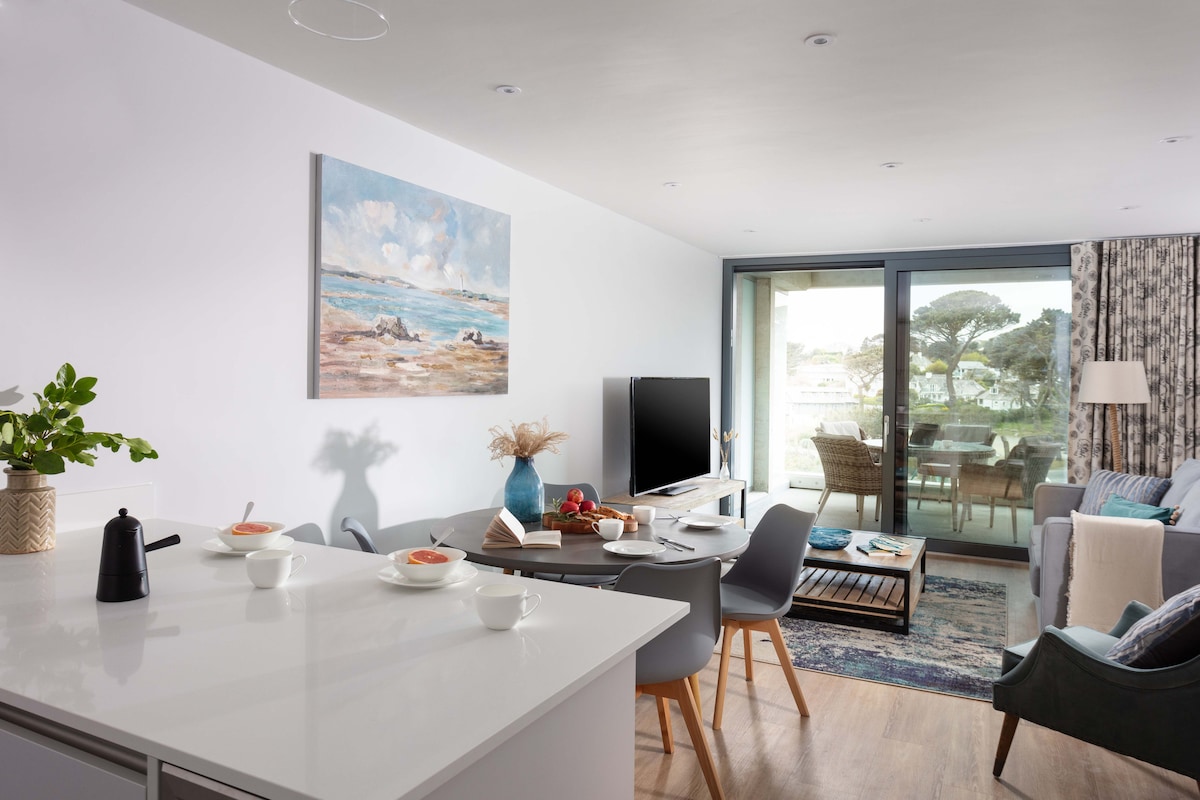 An open-plan living space is depicted, featuring a contemporary kitchen island with a kettle and tableware. A dining area with a round table and grey chairs is visible, alongside a cozy living area with a sofa and coffee table. Floor-to-ceiling sliding doors offer views of the garden and ocean.
