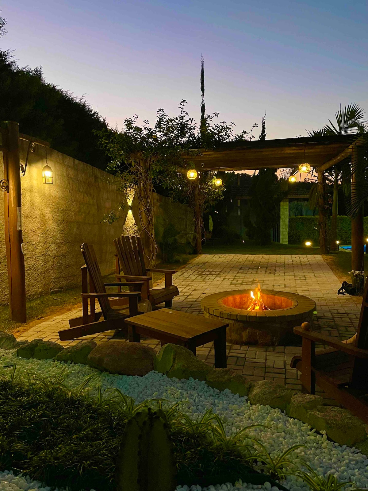An outdoor seating area is present, featuring wooden chairs arranged around a circular stone fire pit. Dim lighting creates a warm ambiance, with greenery surrounding the space. The background showcases a wall with climbing plants, contributing to a tranquil evening environment.