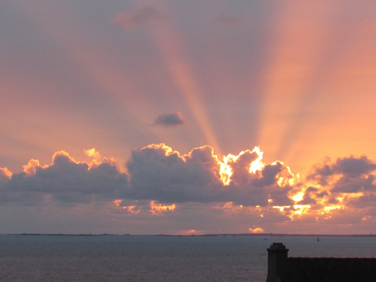 A sunset over the sea is captured, with rays of light breaking through clouds. Soft hues of orange and pink fill the sky, reflecting on the water's surface. The silhouette of a roof is visible in the foreground, contrasting with the vibrant scene above.