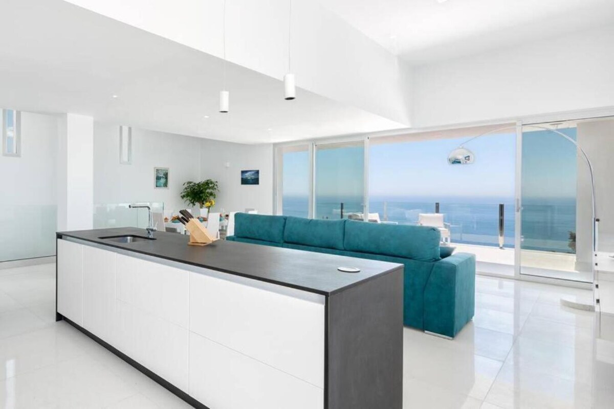 A modern kitchen features a sleek island with a dark countertop and white cabinetry. A spacious turquoise sofa is positioned nearby, offering views through large glass windows that frame the sea and sky. Neutral tones and minimalistic design enhance the open layout.