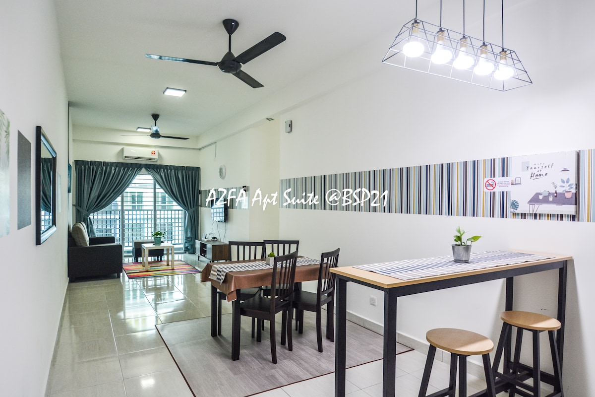 A modern living area is furnished with a dining table and chairs, along with a casual seating space. Large windows allow natural light, while ceiling fans provide ventilation. Decorative wall accents and a small plant enhance the ambiance. The space is well-organized and inviting.