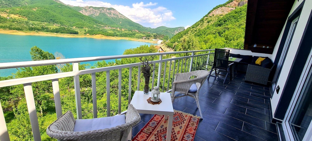 A balcony is shown with a stunning view of a serene lake surrounded by lush green mountains. Two woven chairs and a small white table are comfortably positioned on the tile floor, complemented by a decorative rug and decorative plants.