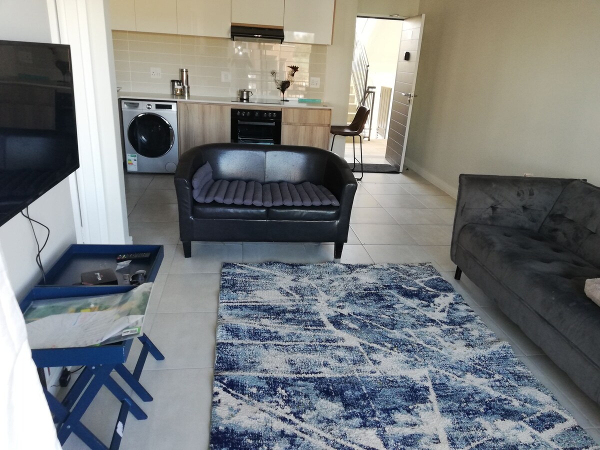 The living area features a cozy black sofa and a gray chaise, positioned on a patterned blue and white rug. An open-plan kitchen with wood accents is visible in the background, alongside a washing machine. Light enters through a nearby sliding door, leading to an outdoor area.