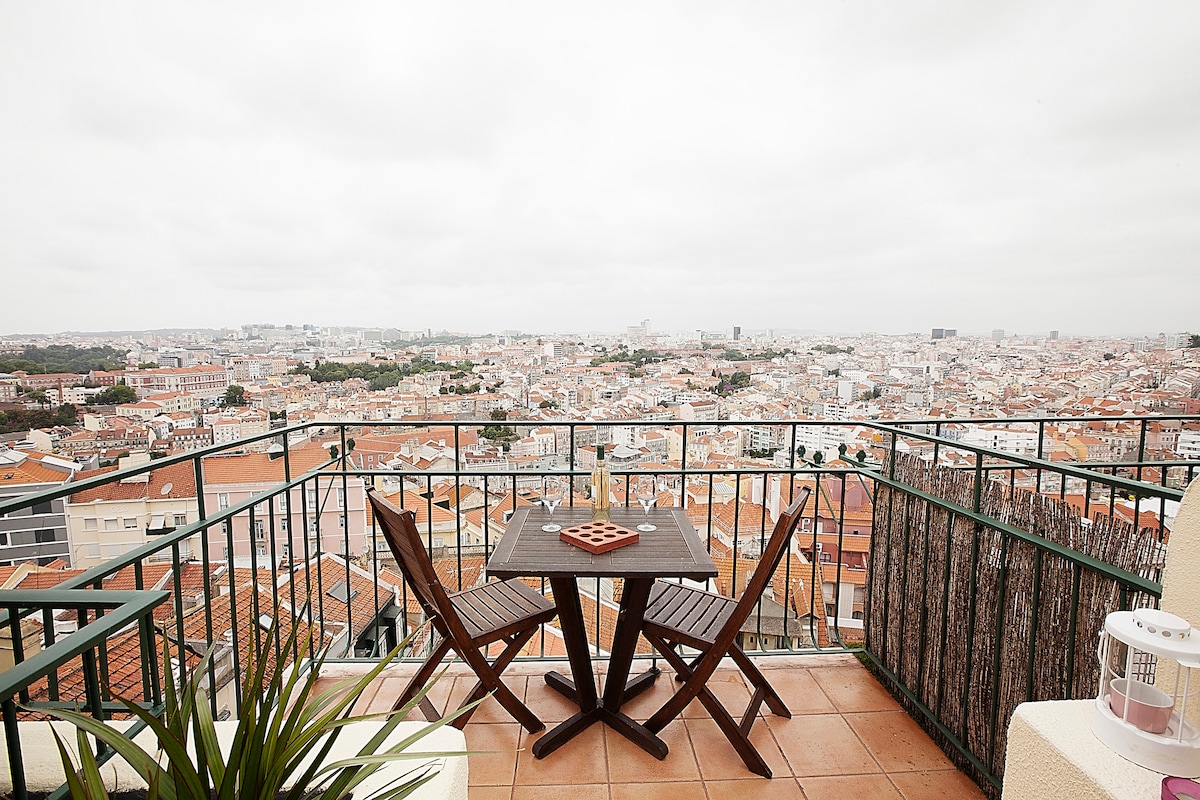 A panoramic terrace is featured with a small wooden table and two folding chairs, overlooking a sweeping view of Lisbon's rooftops. The terrace is surrounded by a decorative railing, with scattered greenery adding a touch of nature to the urban landscape.