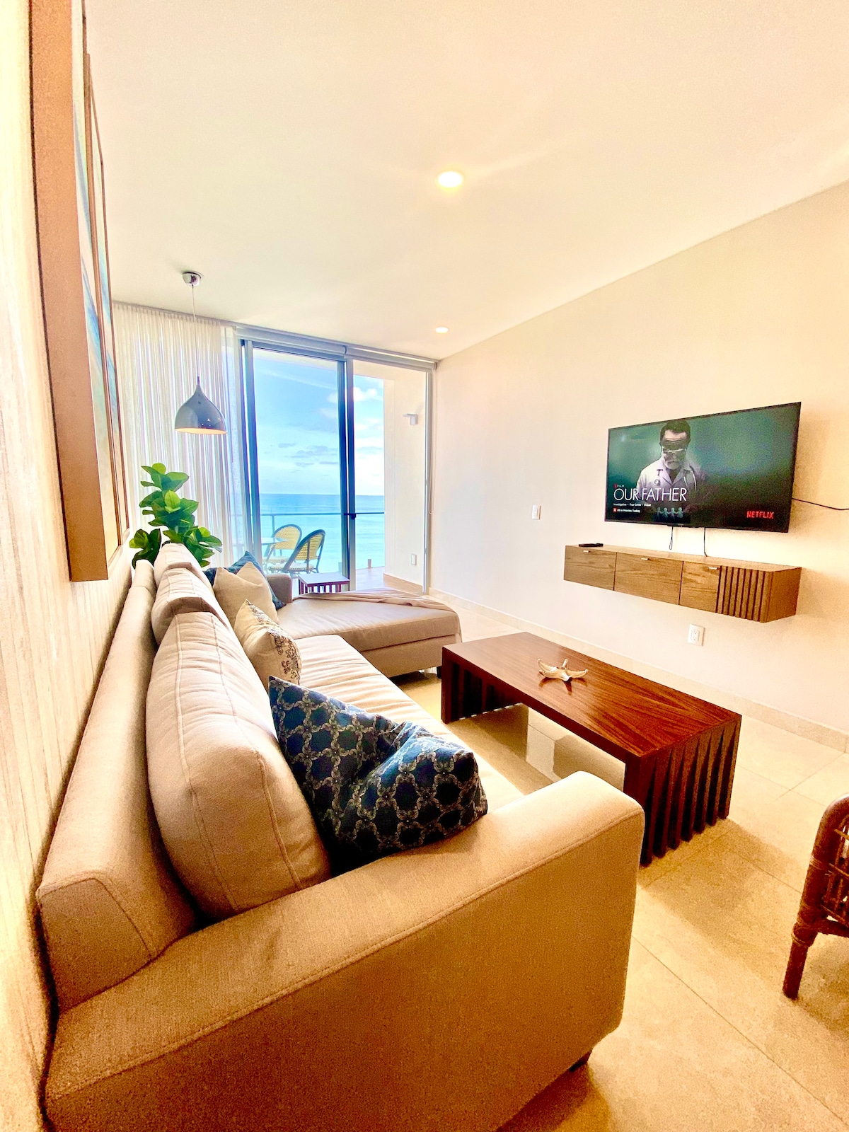 A modern living area features a light-colored sofa adorned with textured cushions, positioned in front of a wall-mounted television. A wooden coffee table sits centrally. Floor-to-ceiling windows allow natural light to fill the space, offering a view of the ocean beyond.