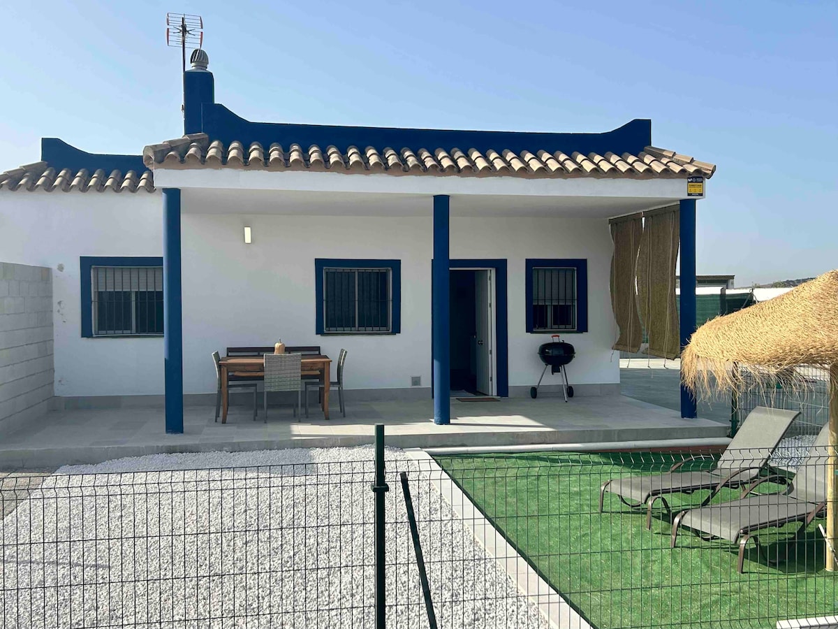 A single-story house features a white exterior with blue accents. The entrance is marked by a spacious porch with a dining table, BBQ grill, and lounge chairs. A small area of artificial grass and gravel surrounds the house, enclosed by a low fence.