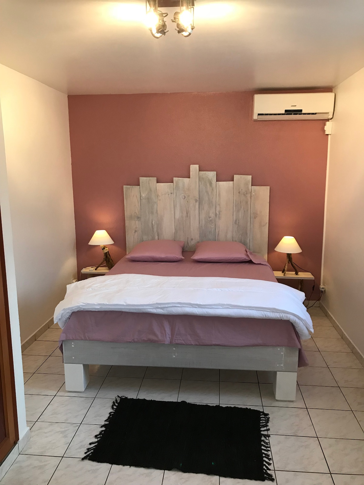 A cozy sleeping area features a spacious bed with light purple bedding and decorative headboard. Two bedside lamps provide soft lighting, and an air conditioning unit is mounted above. Tile flooring adds to the simplicity of the room, complemented by a small dark rug.