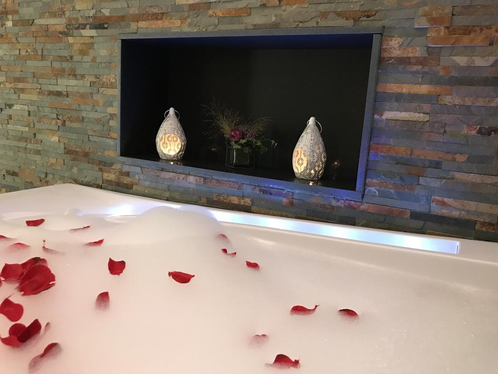 A luxurious bathtub is filled with bubbles, adorned with rose petals floating on top. Behind the tub, decorative lanterns and a floral arrangement are placed on a ledge, creating a serene atmosphere against a textured stone wall.