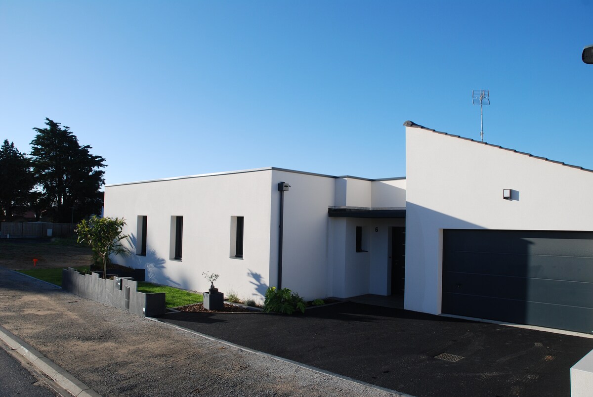 The exterior of a modern home is displayed, featuring a sleek white facade and minimalist design. Large windows allow for natural light. A well-maintained driveway leads to an integrated garage, while landscaped areas with greenery complement the contemporary structure.
