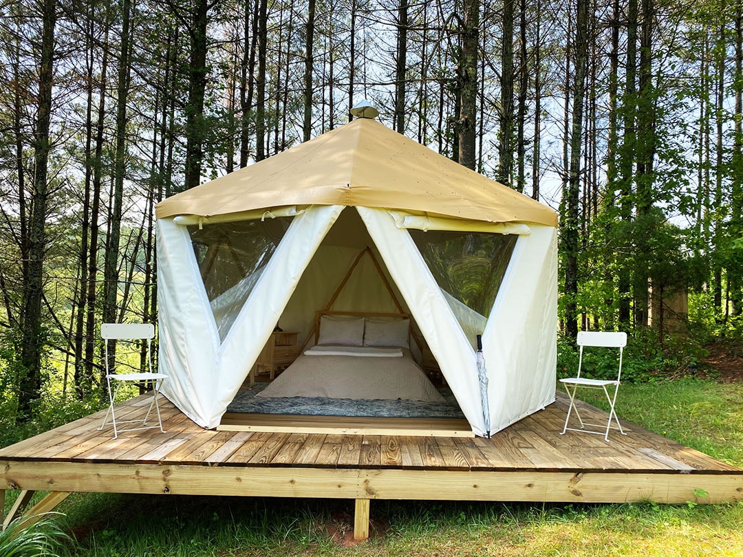 A cozy yurt is nestled among trees, featuring a spacious bed with neutral bedding visible through large, open flaps. Two white chairs are placed on either side of a wooden deck, offering a shaded area to enjoy the surrounding nature.