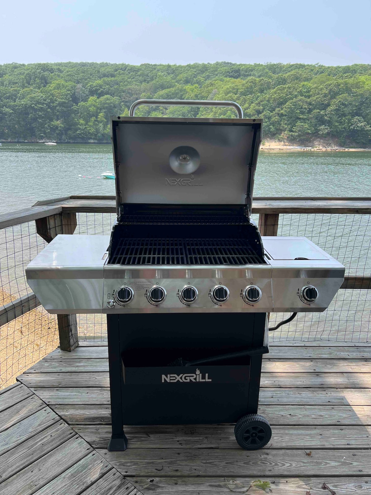 A stainless steel grill is positioned on a wooden deck overlooking the water. The grill's lid is open, revealing multiple grilling grates. Green trees line the background, complementing the calm waterfront setting.