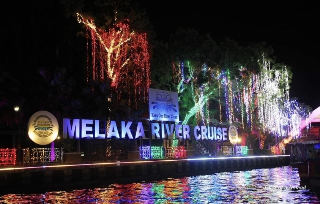 The illuminated Melaka River Cruise sign is brightly displayed along the water's edge, surrounded by colorful fairy lights adorning nearby trees. The scene reflects a festive ambiance, with vibrant colors contrasted against the night sky, creating a captivating view for evening strolls.