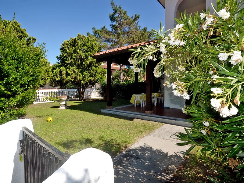 The image features an inviting corner villa with a spacious garden. A covered patio includes a table and chairs under a roof, and vibrant greenery surrounds the area. The path leads to the entrance while a glimpse of the garden reveals well-kept grass and flowering plants.