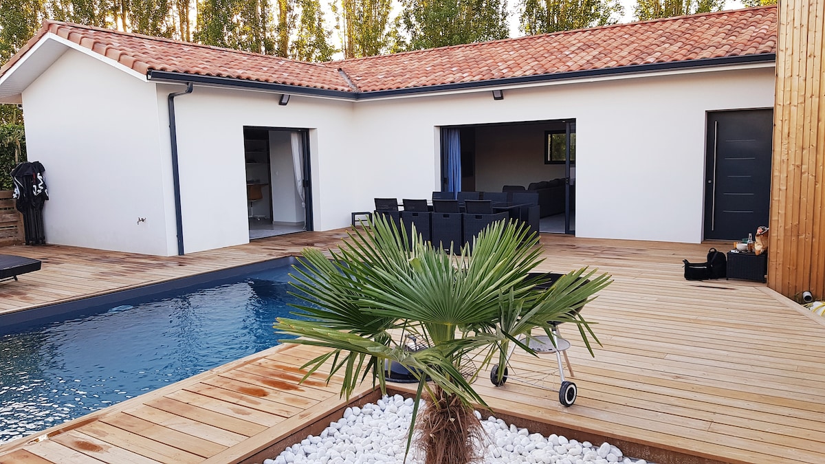 A modern villa's exterior is presented, featuring a wooden deck that surrounds a swimming pool. A lush palm plant adds greenery in the foreground, while sliding glass doors provide access to the spacious living area behind. The roof displays a warm terracotta tile design.