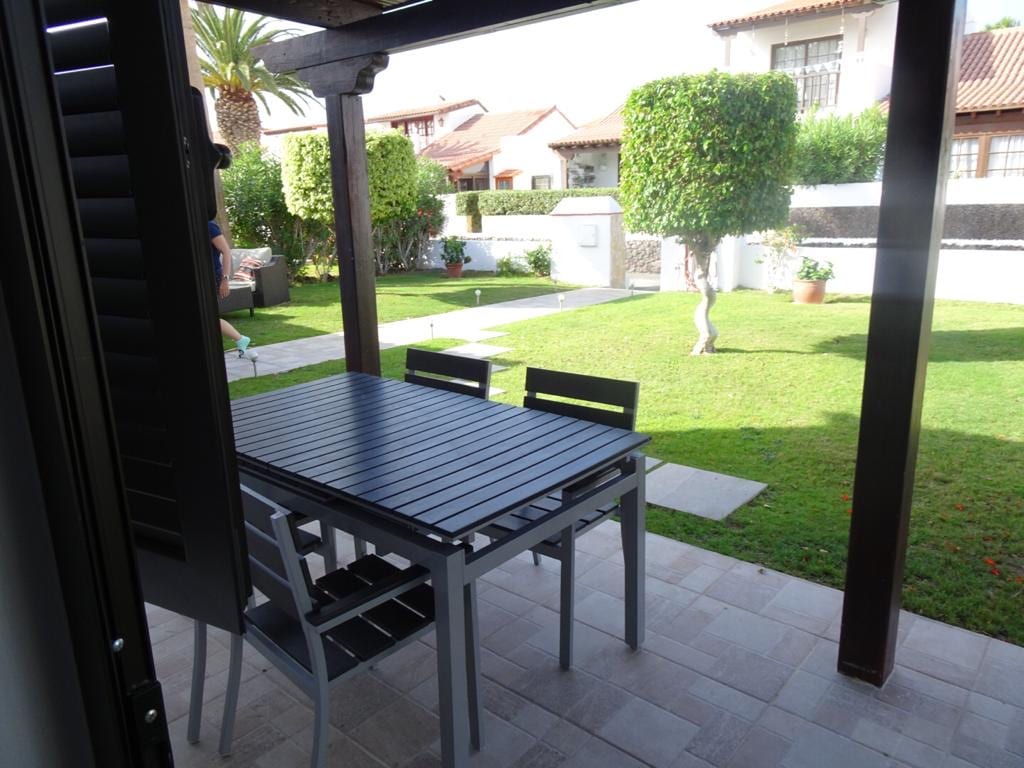 A shaded patio area features a dark wooden table accompanied by several chairs, overlooking a well-maintained garden. Lush greenery and small trees are visible in the background, creating an inviting outdoor space for relaxation.