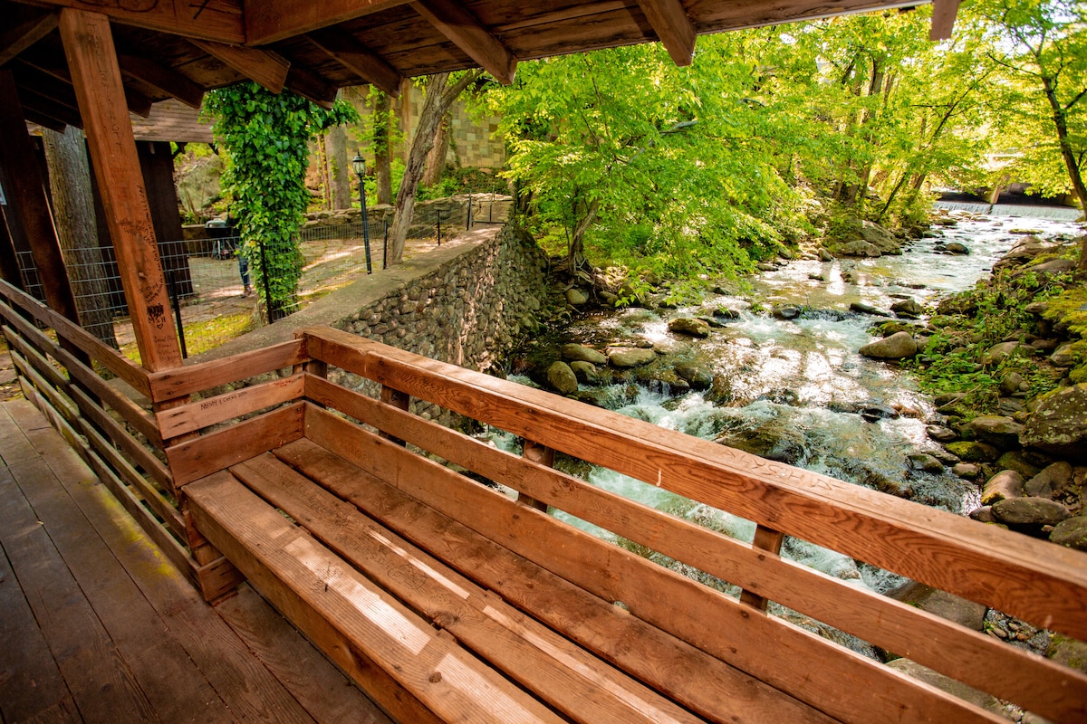 A spacious wooden deck with rustic railings overlooks a gently flowing stream. Lush greenery surrounds the area, providing a serene and natural setting. The soothing sound of the water enhances the tranquil atmosphere, inviting relaxation and enjoyment of the outdoor scenery.