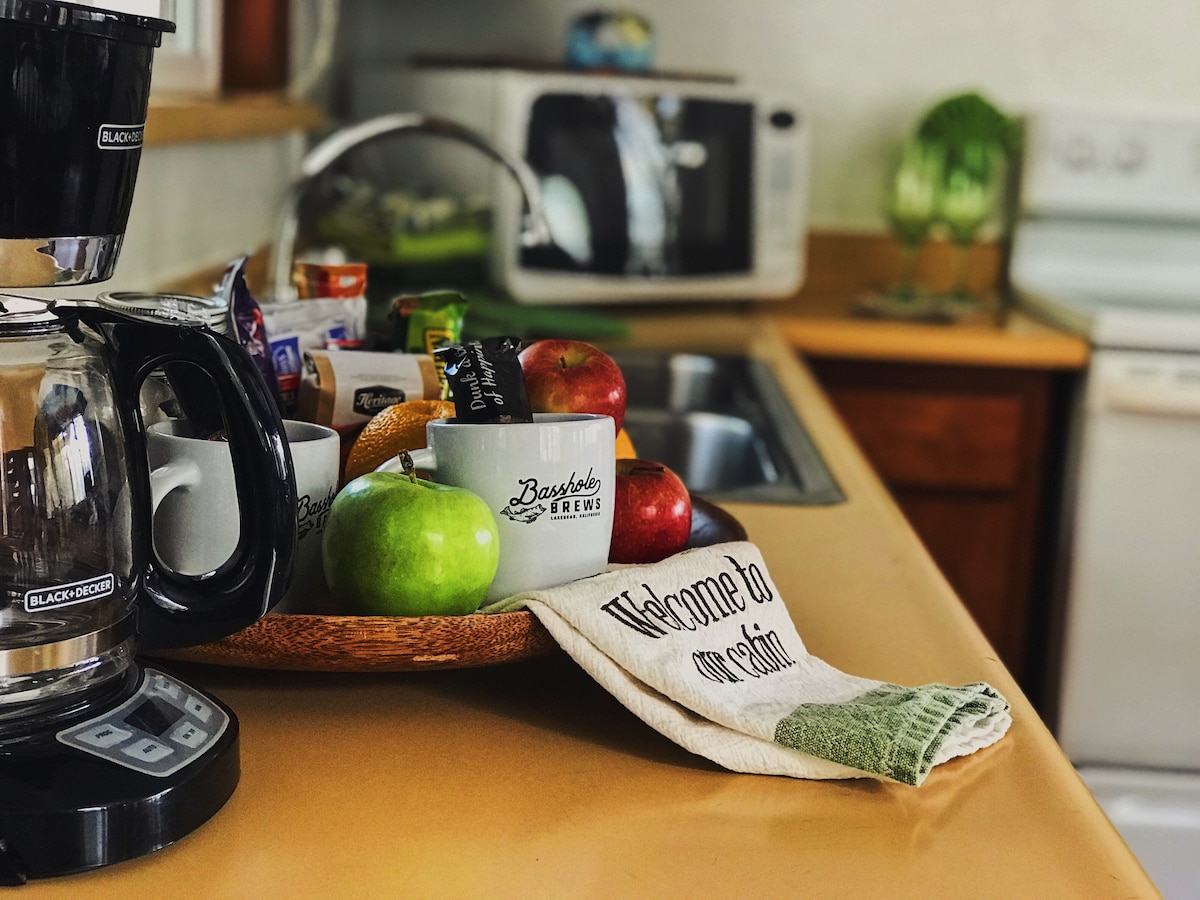 A rustic kitchen area displays a coffee maker next to a tray with mugs, fresh apples, and a dish towel that reads 'Welcome to our cabin.' To the side, a microwave and sink are visible, complementing the warm wooden cabinetry.