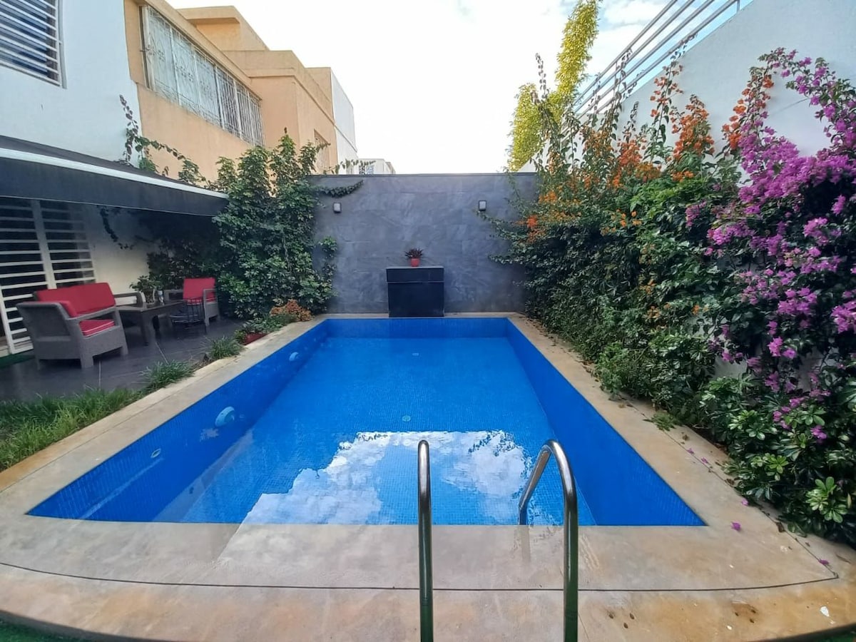 A well-maintained pool is surrounded by lush greenery and colorful flowering plants. Comfortable outdoor seating is visible beside the pool, providing a serene environment for relaxation. The clean water reflects the sky, creating a tranquil vibe in the open outdoor space.
