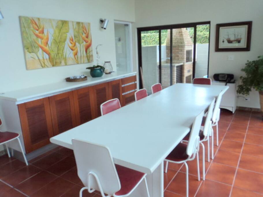 Sala de jantar com mesa para até 12 pessoas, bufê, próxima à churrasqueira e com vista para o jardim.