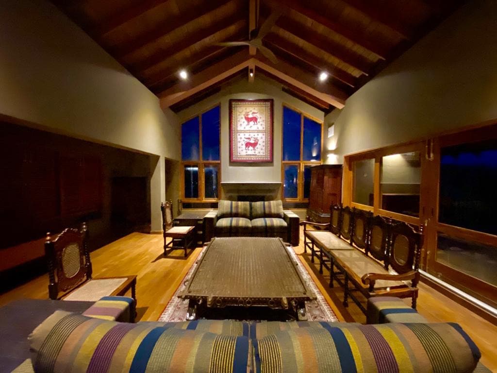 A spacious living area features a high ceiling with wooden beams and large windows that provide ample natural light. Comfortable seating arrangements, including a large sofa and several antique chairs, surround a low table. Decorative wall art adds character to the room.