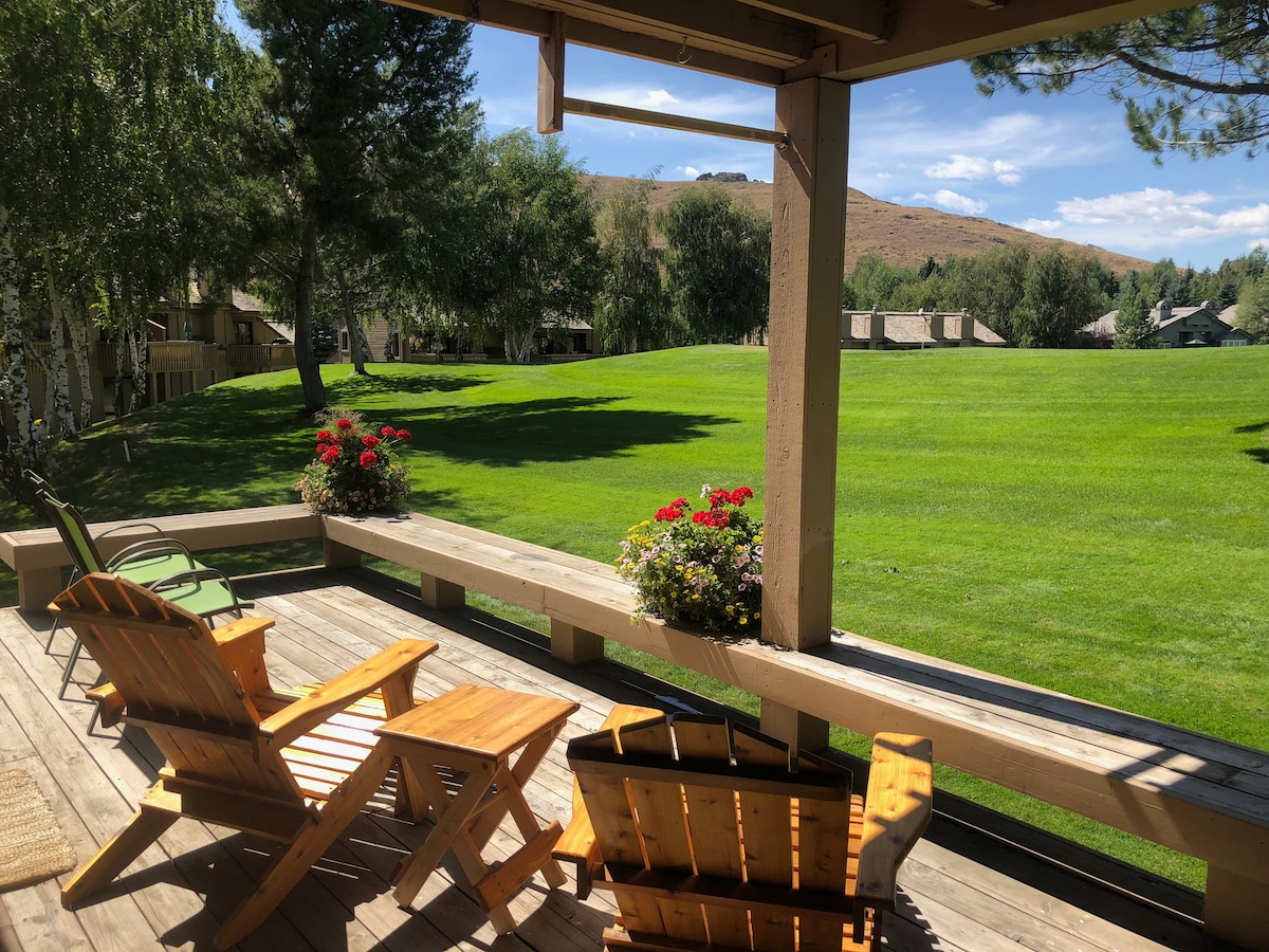 A spacious outdoor deck features wooden lounge chairs alongside potted flowers. The view extends to a well-manicured green lawn, bordered by trees and distant hills under a clear blue sky.