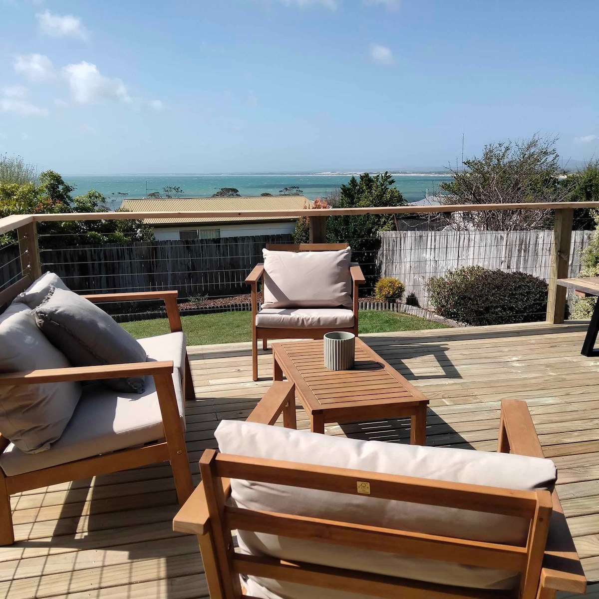 A sun-drenched deck features comfortable outdoor seating with plush cushions, positioned to offer panoramic ocean views. A wooden coffee table complements the seating area, while gentle greenery and the coastline are visible in the background, creating an inviting space for relaxation.