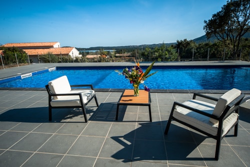 A tranquil outdoor area is showcased, featuring a large blue swimming pool surrounded by a tiled patio. Two chairs are positioned beside a table, adorned with fresh flowers. Lush greenery and distant hills are visible in the background under a clear blue sky.
