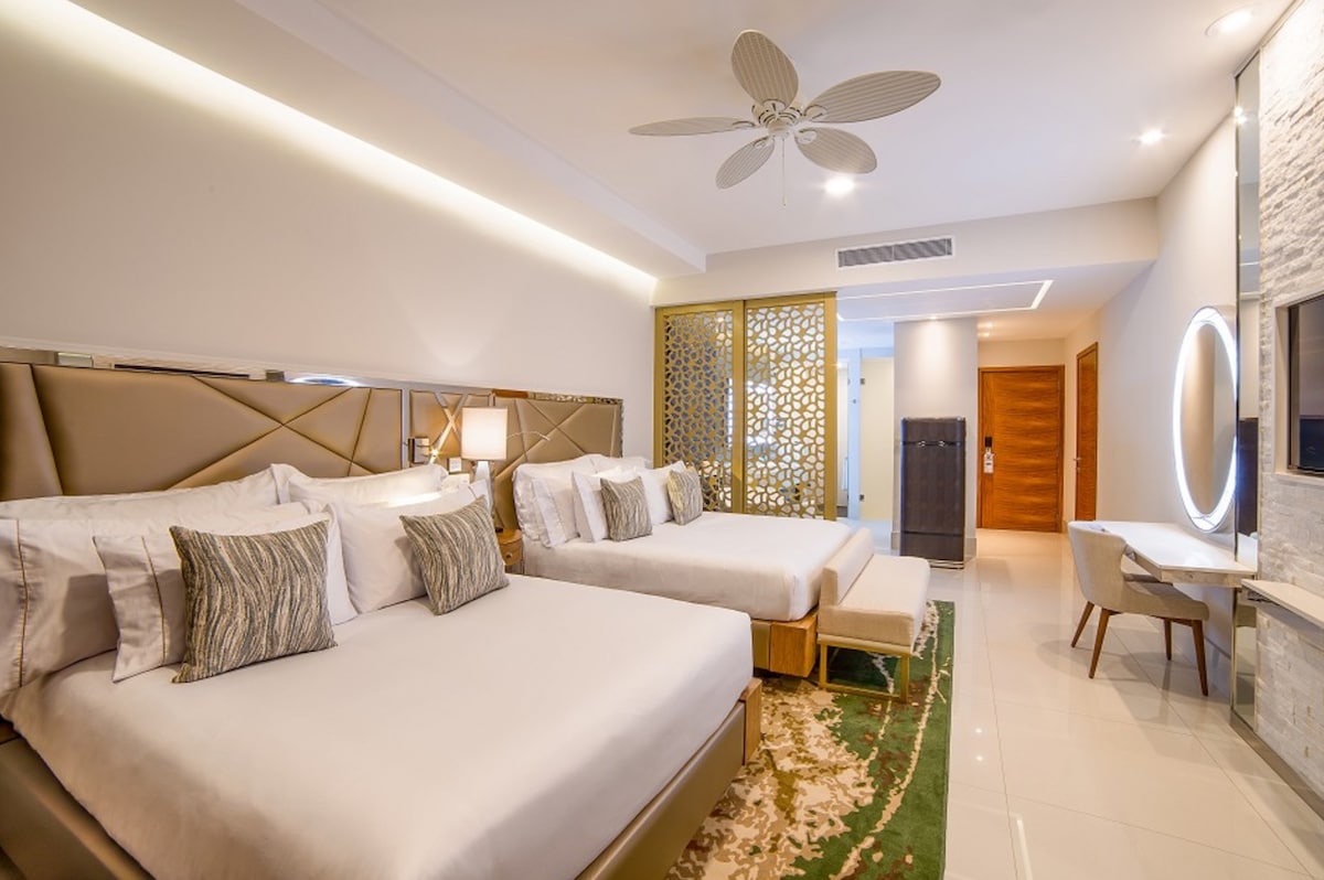 The spacious sleeping area features two large beds with decorative pillows. Soft lighting is provided by a ceiling fan and wall sconces. A decorative partition adds privacy, while a modern desk and TV unit are positioned along a light-colored wall. A rug adds warmth to the tiled floor.