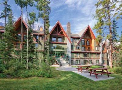 The exterior of a multi-story building is surrounded by trees and green grass. The design features triangular roof peaks and large windows. A picnic area with red benches is visible on the lawn, providing a space for relaxation outdoors.