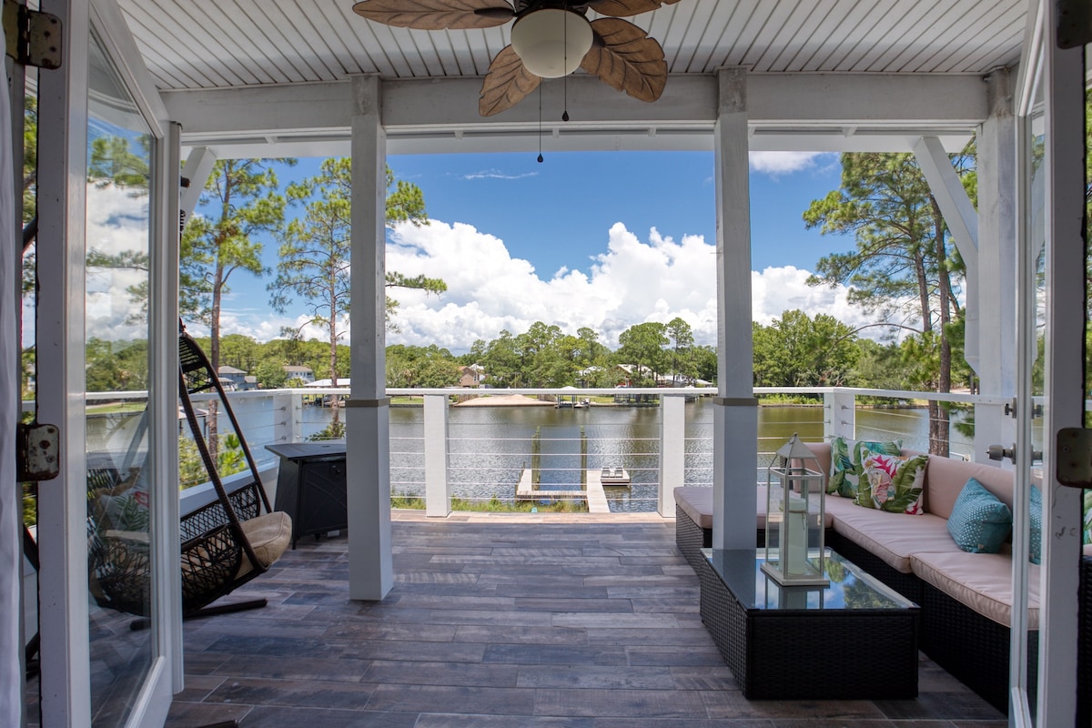 A spacious screened-in porch features comfortable seating with decorative cushions, looking out over a serene waterway. Tall trees and clouds are visible in the background, while the porch flooring consists of tile, adding a modern touch to the relaxed outdoor environment.