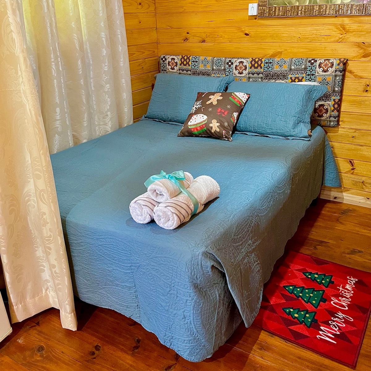 A cozy sleeping area is presented with a spacious bed covered in a blue quilt. Two decorative pillows are arranged on top. Fresh towels are neatly rolled and placed on the bed. A festive holiday mat features a Christmas tree design near the bed.