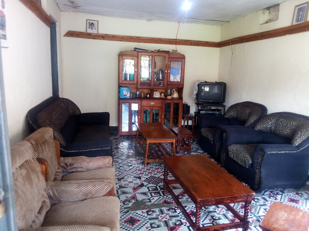 A cozy sitting room features a mix of sofas in various patterns, complemented by wooden coffee tables. An ornate cabinet holds decorative items, while a television is positioned on a stand. The floor is adorned with a patterned rug, contributing to a homey atmosphere.