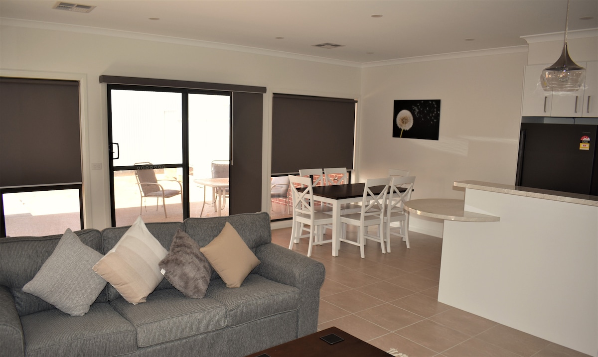 A spacious living area features a comfortable grey sofa adorned with neutral cushions. Natural light enters through large glass doors leading to an outdoor area. A dining space includes a white table and chairs, positioned adjacent to a modern kitchen with sleek cabinetry.