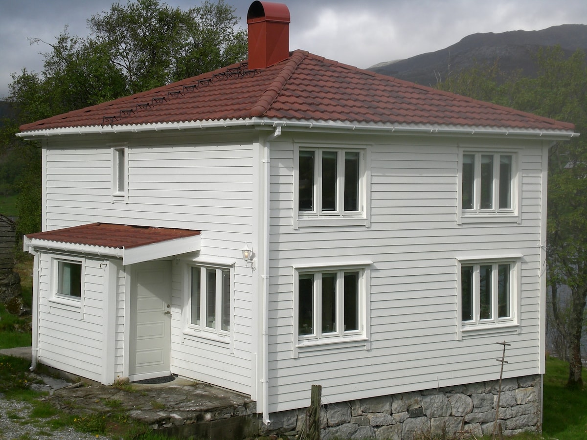 A two-story country house is showcased, featuring a white wooden exterior with a red-tiled roof. Multiple large windows allow natural light to enter the space. A small overhang provides shelter at the entrance, while stone elements form the foundation, complementing the serene surrounding landscape.