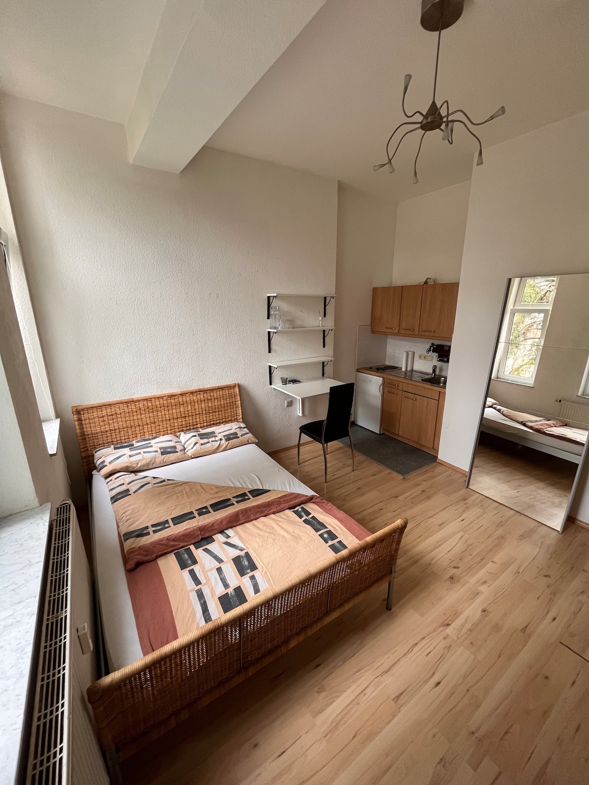 A bright room features a spacious bed with a woven headboard and colorful bedding. Nearby, a compact kitchenette is equipped with wooden cabinets. A mirror reflects the room's layout, and light enters through a large window, enhancing the inviting space.