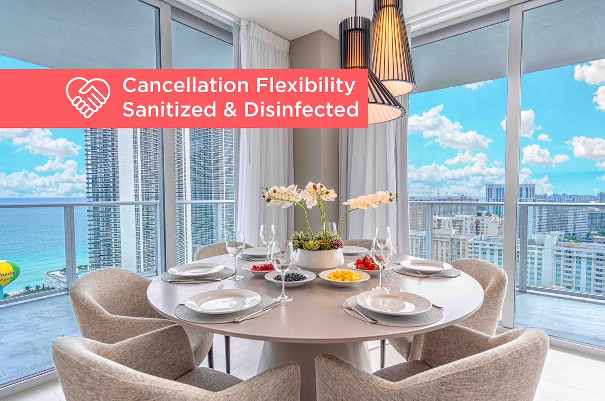 A dining area is presented with a round table set for four, featuring elegant dishware and a centerpiece of orchids. Floor-to-ceiling windows showcase expansive views of the ocean and city skyline, filling the space with natural light.