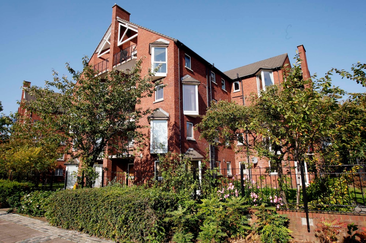 The apartment complex features a charming red brick exterior, set amidst landscaped gardens. Lush greenery and flowering plants frame the property, while a clear blue sky extends overhead, enhancing the inviting appearance of this residential area.