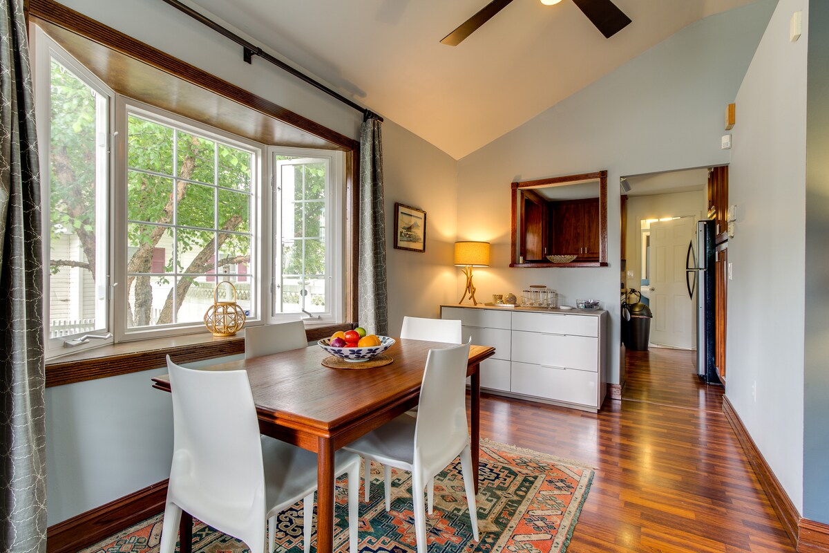 A dining area features a wooden table surrounded by four white chairs. Natural light filters through a large window, illuminating the space. The room includes a well-furnished sideboard and a view into the adjoining kitchen, with a ceiling fan enhancing air circulation.