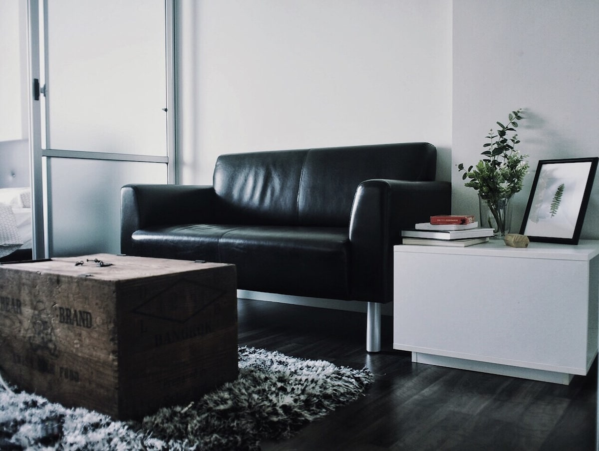 A modern living area features a sleek black sofa positioned beside a minimalist coffee table made of wood. A small decorative plant and a framed picture add a touch of greenery. Soft, textured rug enhances the comfort of the space.